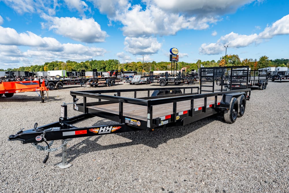 Black dual-axle flatbed trailer with wooden deck and side rails on a gravel lot at a trailer dealership