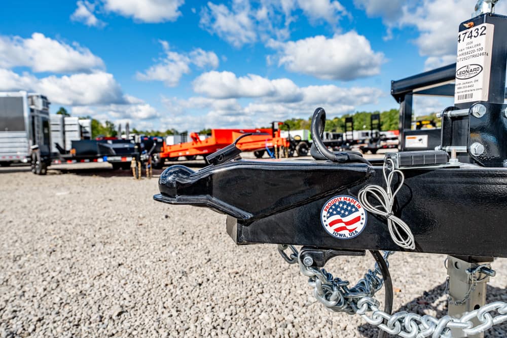 Close-up of a black trailer hitch with a coupler and safety chains in a gravel lot, featuring a 'Proudly Made in USA' sticker on the hitch edge with other trailers in the background.