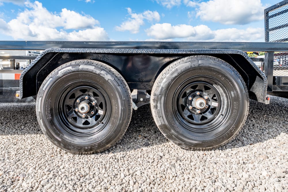 Dual-axle trailer wheels with black rims on a gravel surface under a metal trailer frame, outdoors under a blue sky.