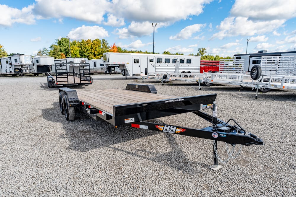 Black dual-axle flatbed trailer with a wooden deck parked on a gravel lot among other horse trailers.