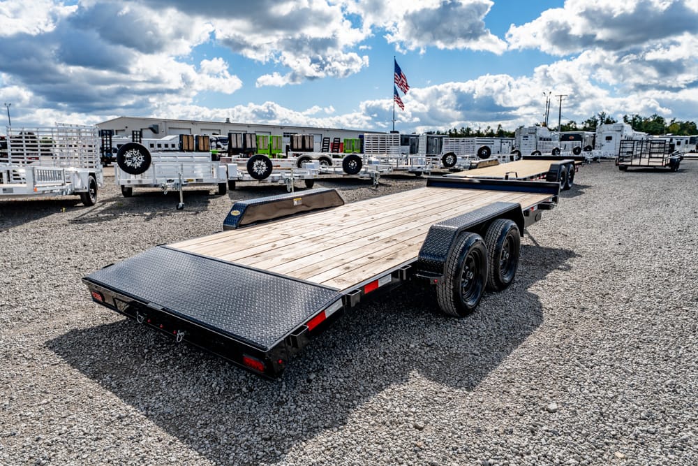 Flatbed car trailer with dual axles on a gravel lot, surrounded by other trailers and an American flag in the background.