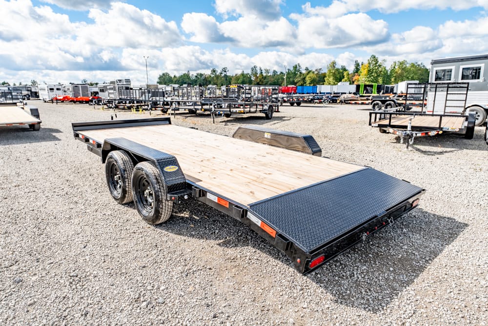 Dual-axle flatbed trailer with a wooden deck and diamond-plate ramp, parked in a gravel lot with other trailers in the background.