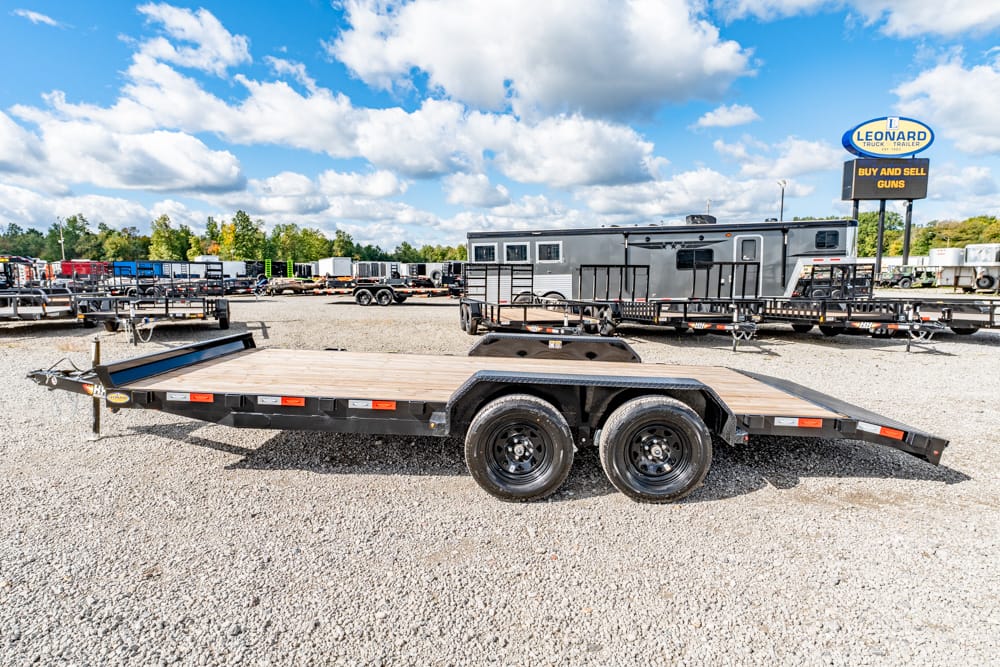 Black dual-axle flatbed trailer with a wooden deck sits on a gravel lot among other trailers and rigs in the background.