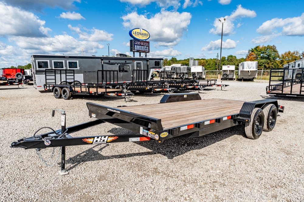 Flatbed trailer with wooden deck and dual rear wheels parked on a gravel lot, with other trailers visible in the background.