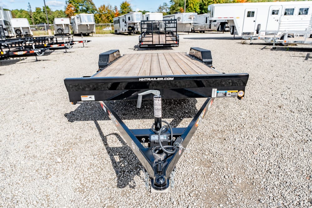 Front view of a black flatbed trailer with a wooden deck and A-frame hitch, parked on a gravel lot with other trailers and RVs in the background.