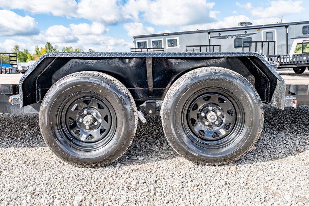 Black steel dual-axle trailer with two large tires on a gravel lot; a silver RV sits in the background behind the trailer edge.