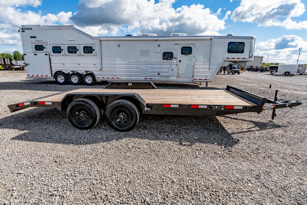 Flatbed trailer with a wooden deck and tandem wheels parked on a gravel lot, with a large white fifth-wheel RV in the background.