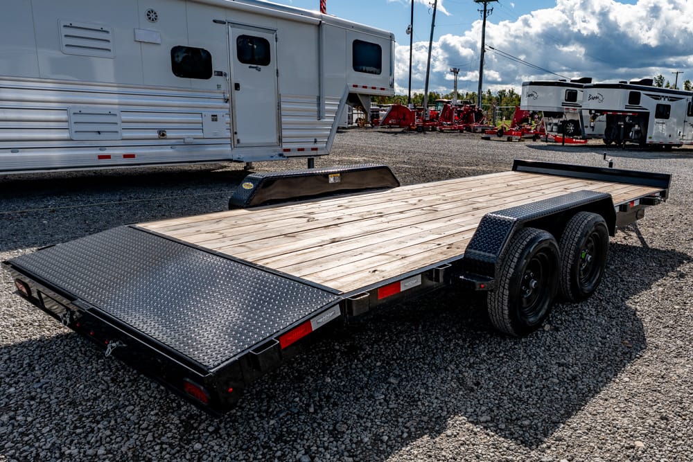 Flatbed trailer with a wooden deck and dual-axle wheels on a gravel lot, with large RVs in the background.