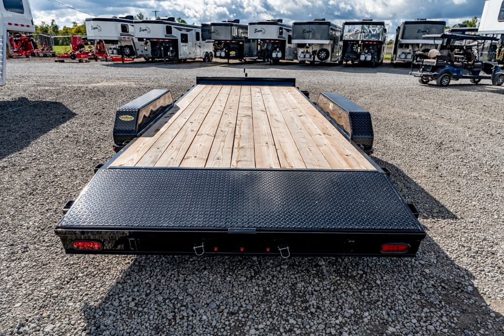 Flatbed trailer with a wooden deck and diamond-plate fenders sits on a gravel lot, with several RVs and trailers in the background.