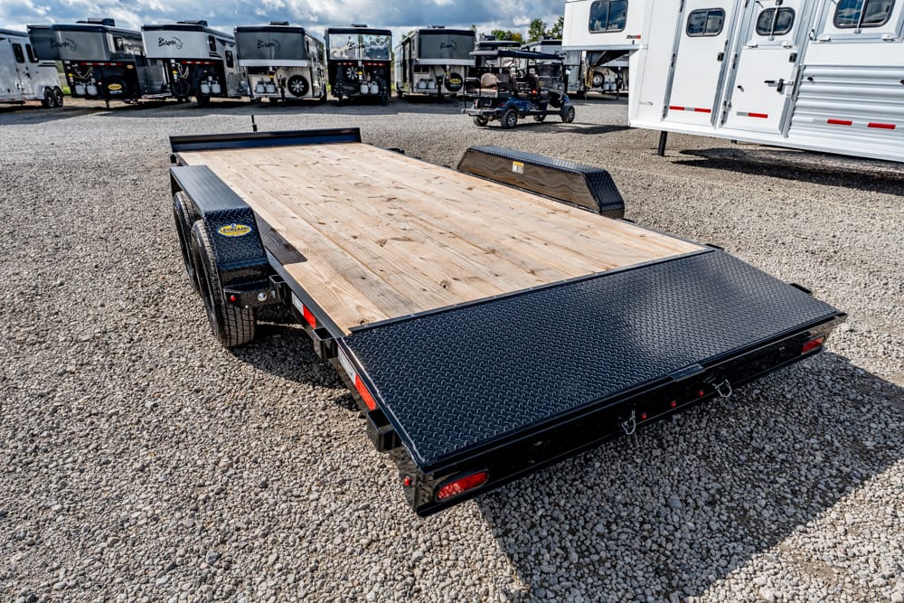 Black dual-axle flatbed trailer with a wooden deck and diamond-plate rear ramps, parked on a gravel lot among other trailers