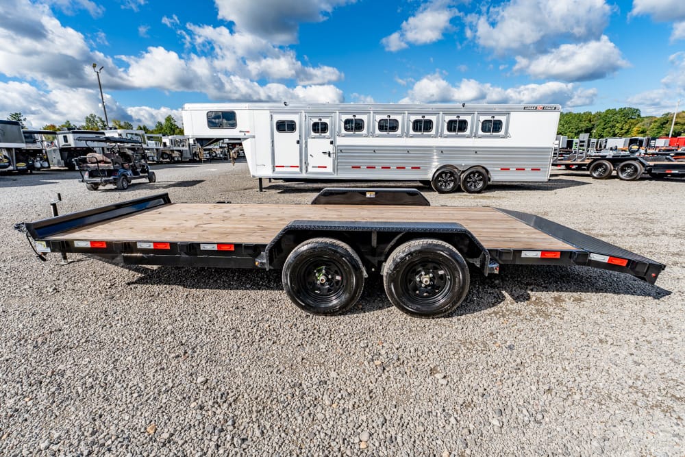 Black dual-axle flatbed trailer with a wooden deck on a gravel lot, with a white horse trailer in the background under a blue sky.