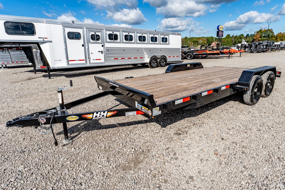 Dual-axle flatbed trailer with a wood deck sits on a gravel lot, with a horse trailer and other trailers in the background.