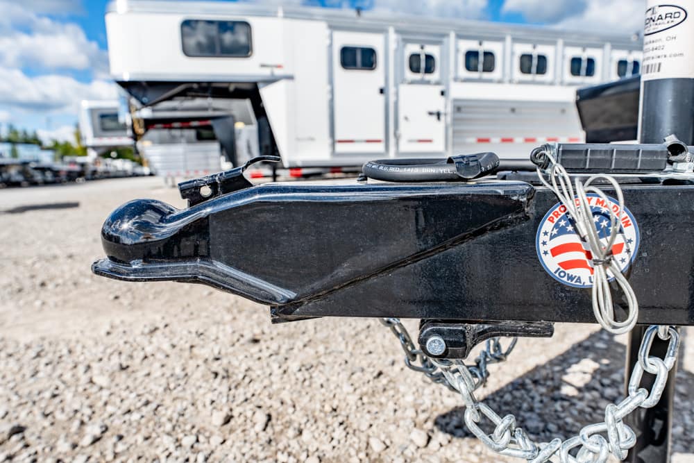 Close-up of a black trailer hitch with safety chains and a circular badge, in a gravel lot with white trailer trailers in the background.