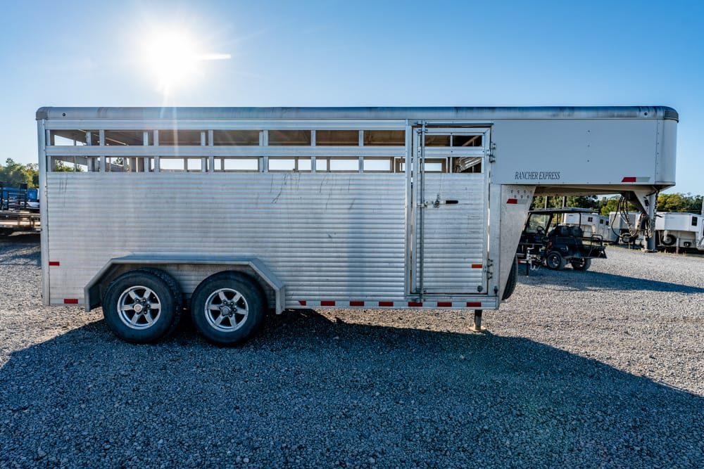 Silver livestock trailer with dual axles parked on a gravel lot under a bright sun.