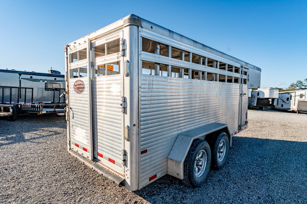 Silver aluminum two-axle horse trailer parked on a gravel lot, with other trailers in the background.