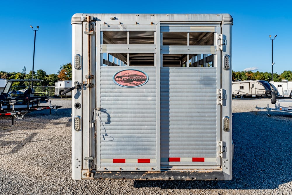 Rear view of a silver aluminum horse trailer with its double doors closed, a central logo plaque, and red reflective strips on a gravel lot under a blue sky.