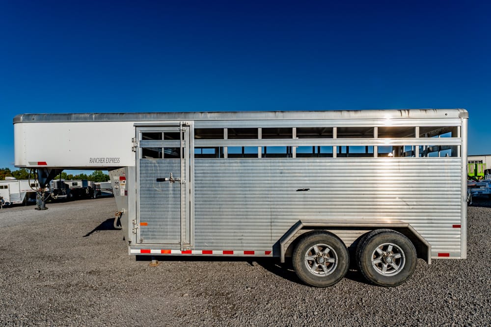 Silver aluminum livestock trailer with vented side panels parked on a gravel lot under a clear blue sky, labeled 'RANCHER EXPRESS' at the front.