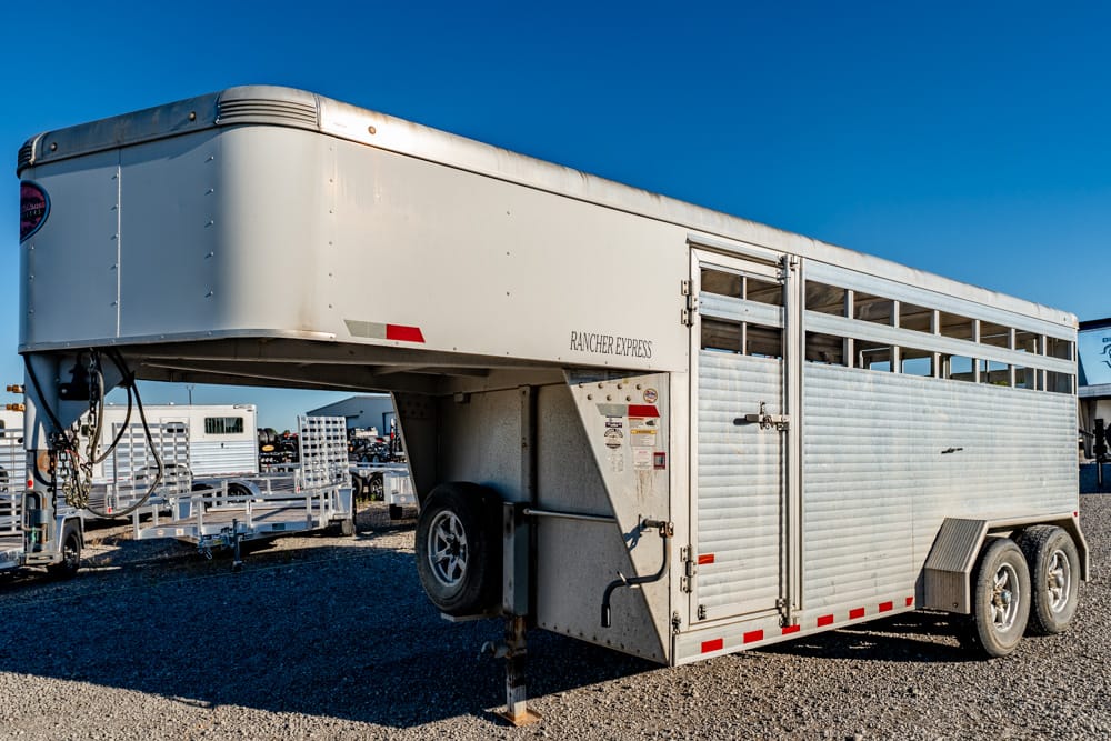 White gooseneck livestock trailer (Rancher Express) parked on a gravel lot, with two axles and slatted side vents on the right side of the trailer.”,
