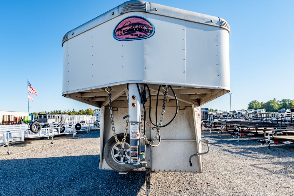 Front view of a white Sundowner gooseneck trailer in a gravel lot, with chains and hitch in the foreground and other trailers in the background.