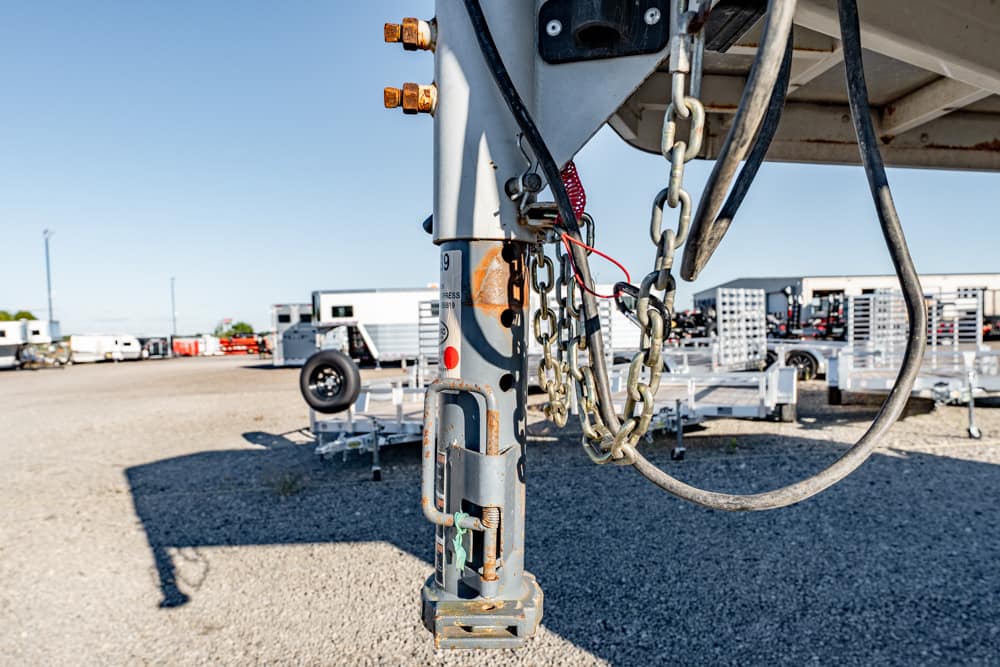 Close-up of a trailer hitch with safety chains and wiring in a gravel trailer yard.
