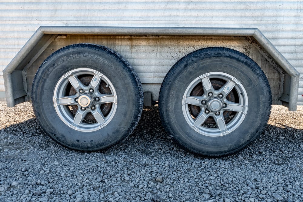 Two trailer wheels with silver rims sit on a gravel surface beside a corrugated metal trailer body.