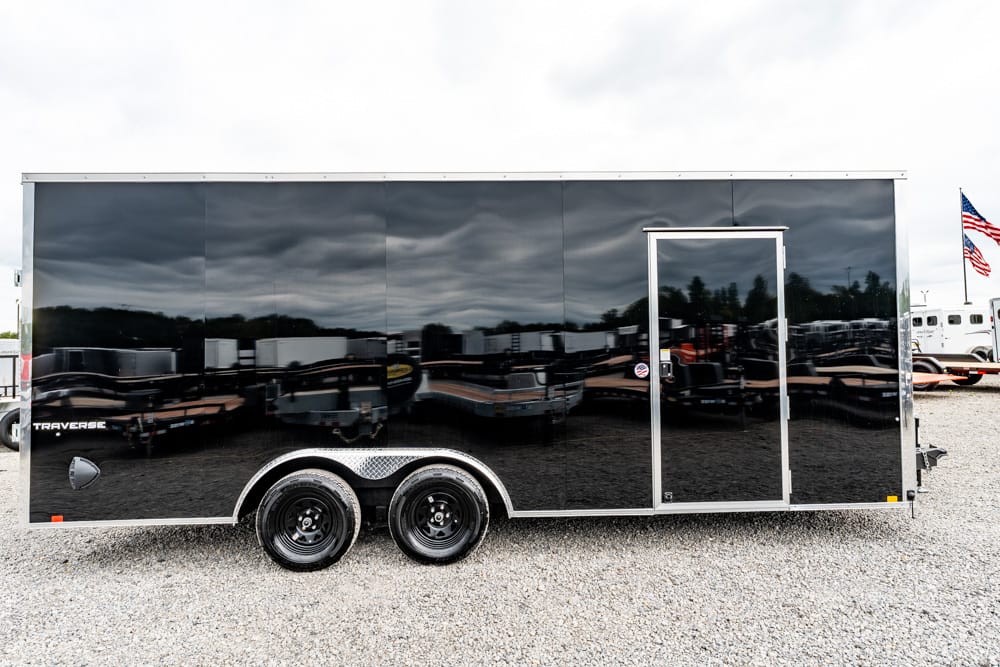 Black enclosed trailer with a single entry door and two-axle wheels, parked on gravel; its glossy surface reflects nearby trailers and an overcast sky.