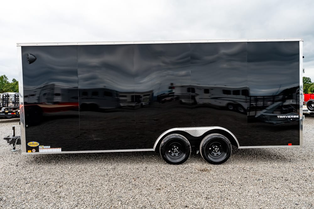 Black enclosed cargo trailer with dual axles on a gravel lot, reflective surface showing surrounding vehicles and structures.
