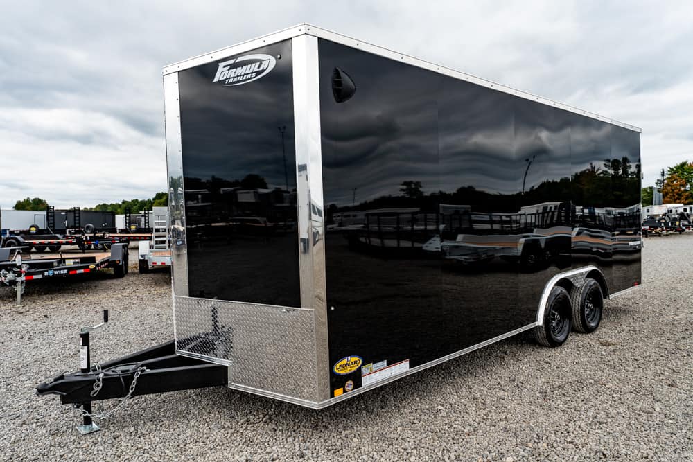 Black enclosed cargo trailer with tandem axles and chrome diamond-plate trim in a gravel lot at a trailer dealership, reflecting surroundings on its glossy panels.