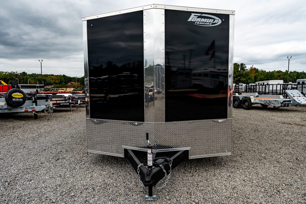 Front view of a black enclosed cargo trailer with diamond-plate front, hitch and chains on a gravel lot.