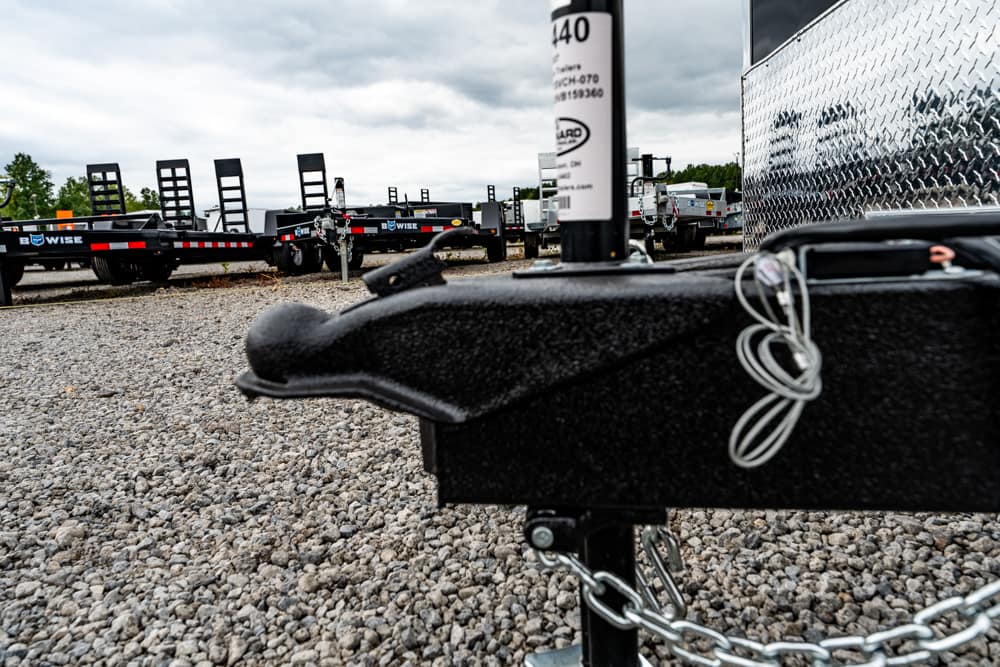 Close-up of a black trailer hitch on a gravel lot, with safety chains looped and diamond-plate trailer to the side.