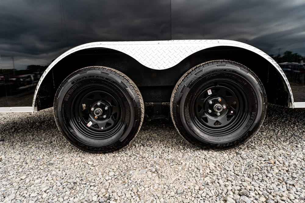 Dual axles on a black trailer with diamond-plate fender over the wheels on a gravel surface at a truck yard