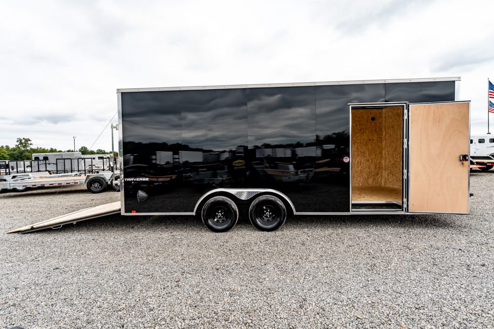 Black enclosed trailer on a gravel lot with the rear ramp lowered and the side door open, exposing a wood-lit interior and tandem wheels