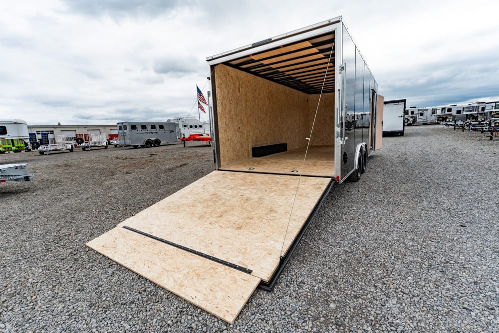 Open enclosed cargo trailer with a wooden loading ramp extended onto a gravel lot, ready for loading.