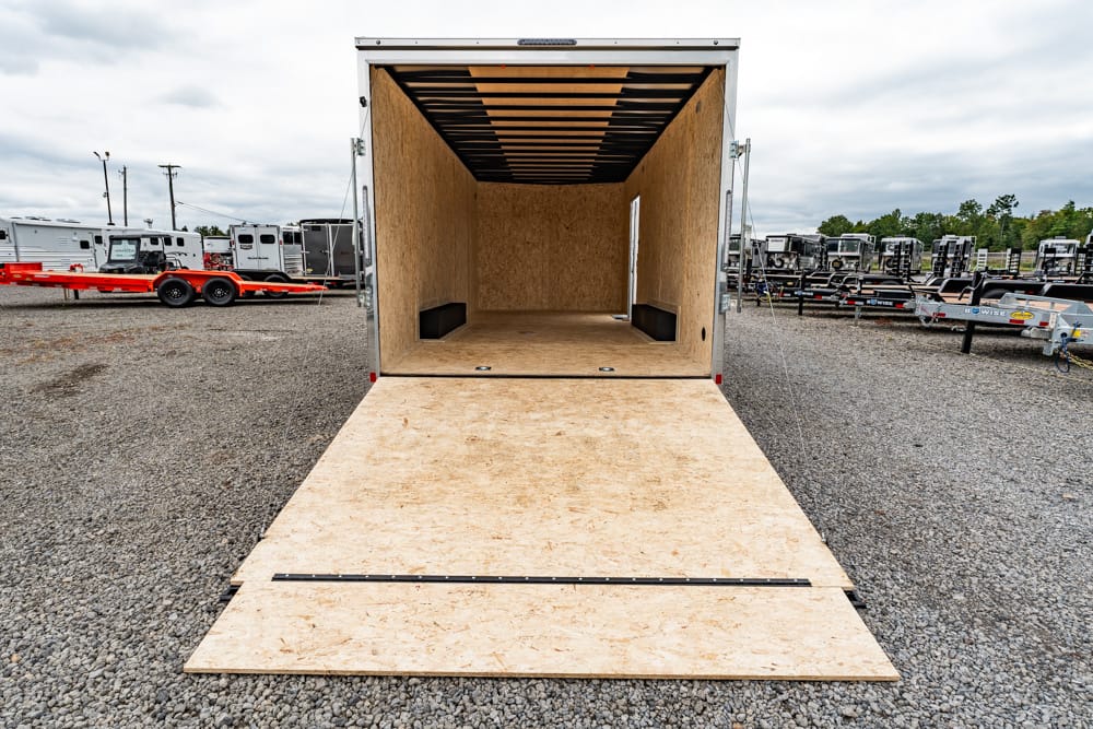 Open cargo trailer with plywood interior and ramp lowered, on a gravel lot with other trailers in the background.