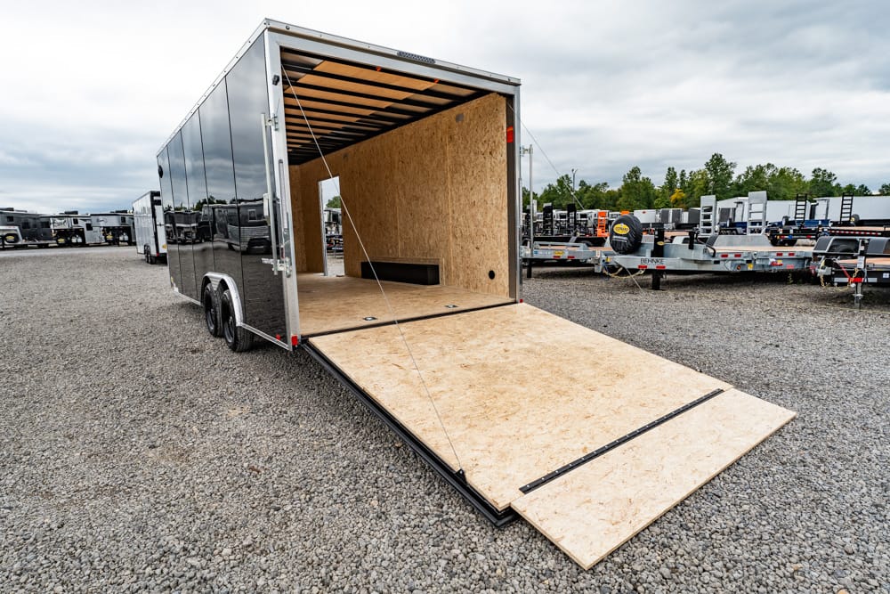 Enclosed cargo trailer with its plywood ramp lowered for loading, parked on a gravel lot with other trailers in the background.