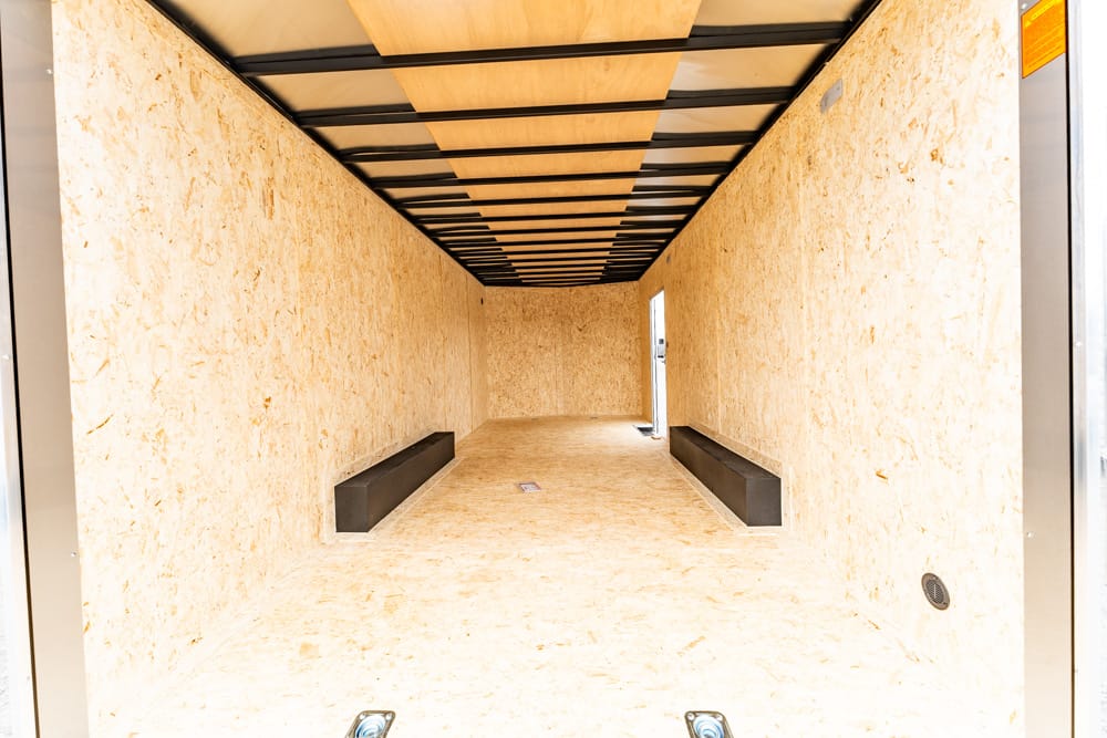 Empty enclosed trailer interior with light wood OSB walls, a wood floor, and black benches along both sides leading to a door at the far end.