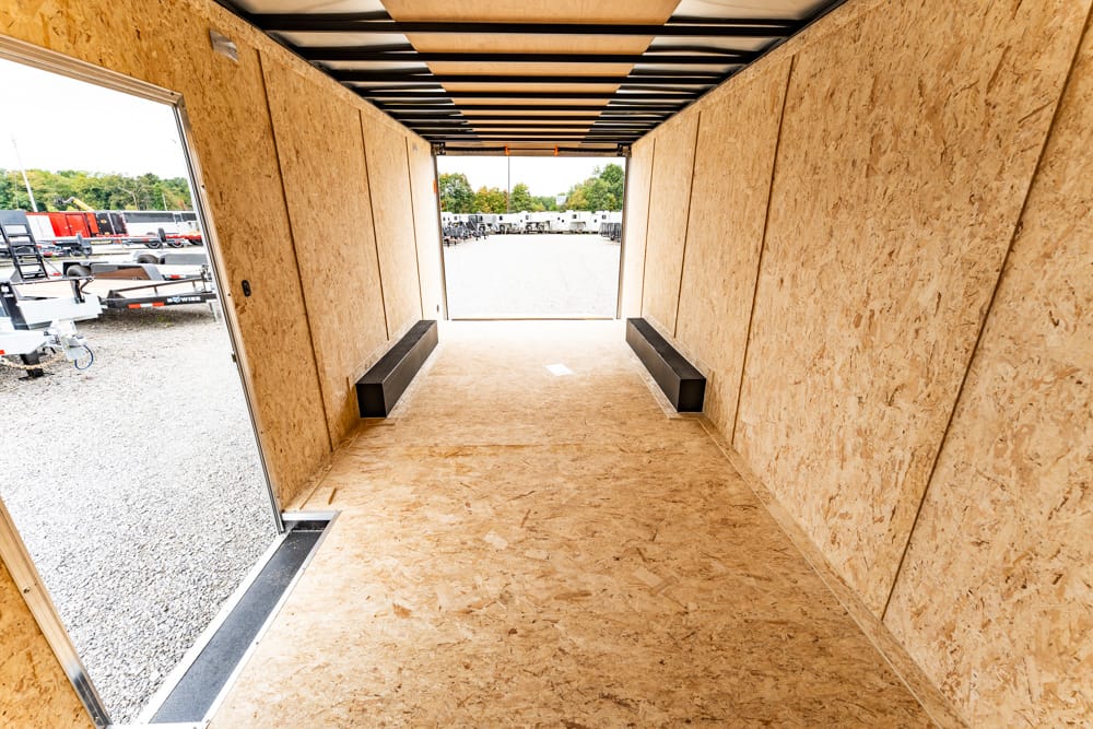 Interior view of an empty plywood-walled trailer with two black benches along the sides, looking toward the open back door to a gravel lot.