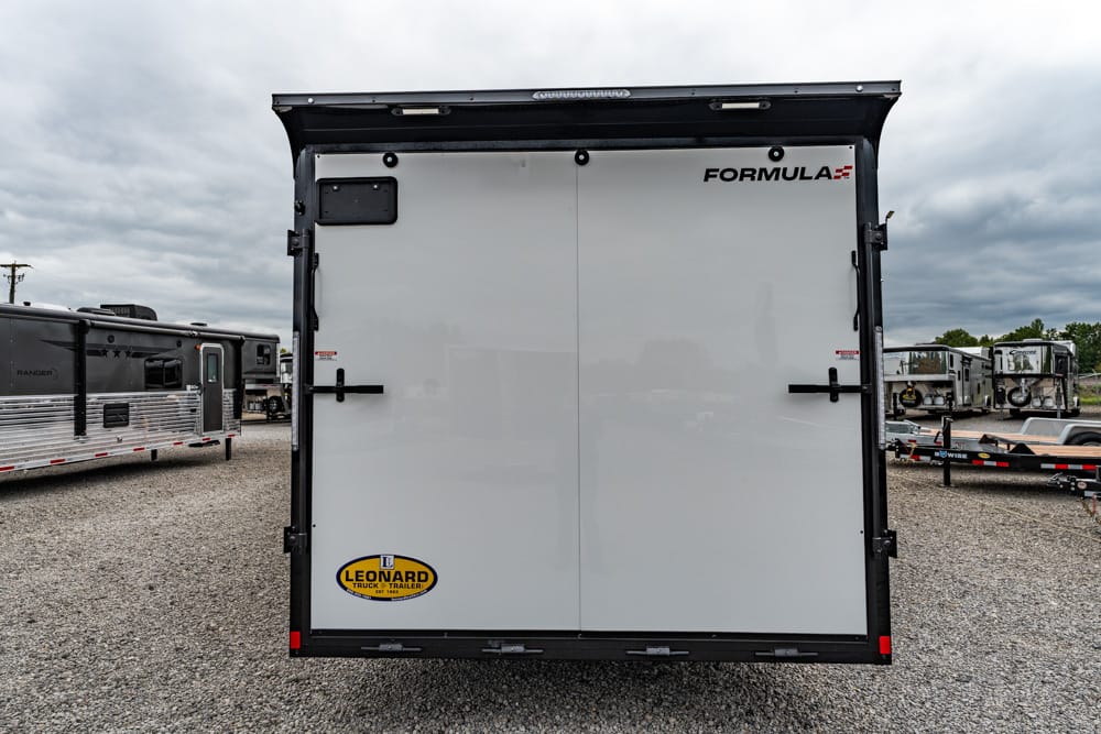 Rear view of a white enclosed cargo trailer with double doors, branded Formula, parked on a gravel lot with other trailers nearby.