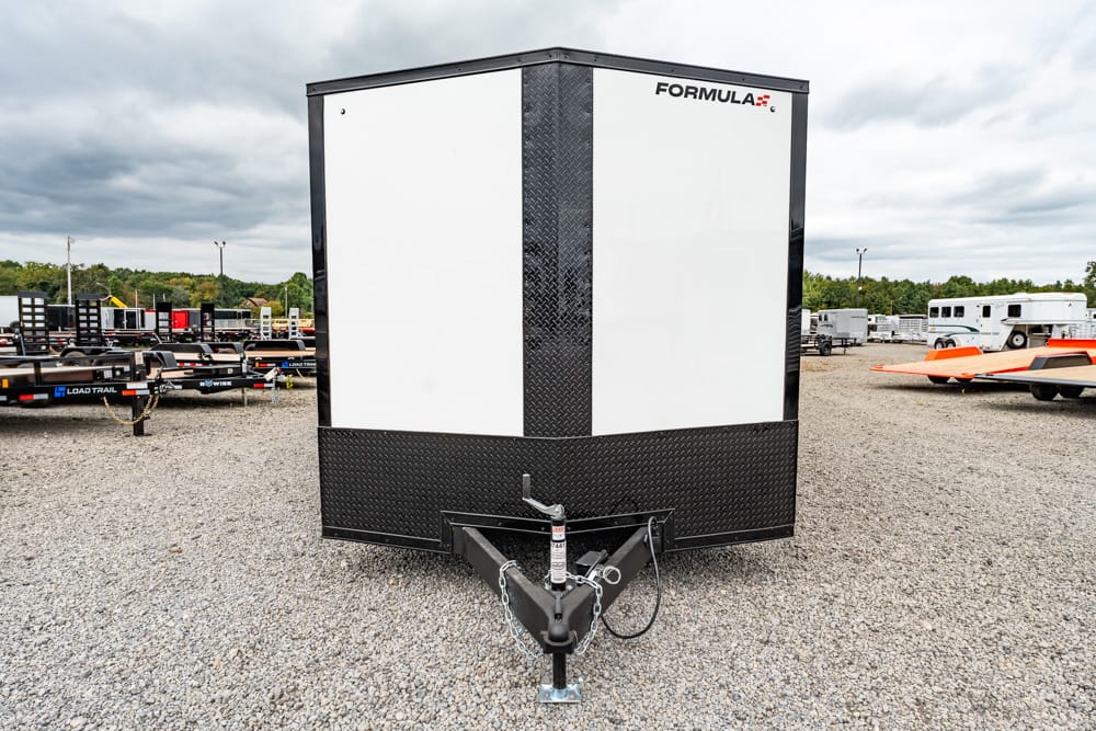 Front view of a black-and-white enclosed cargo trailer with a diamond-plate front, branded Formula, hitched on a gravel lot with other trailers in the background.