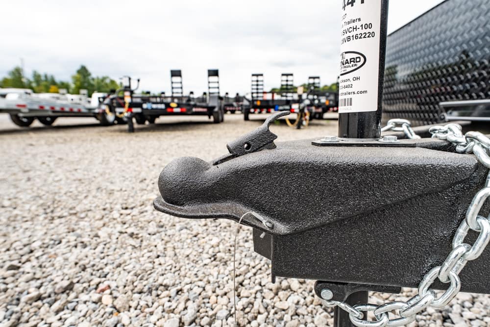 Close-up of a trailer hitch with a safety chain on a gravel lot; row of trailers in the background.