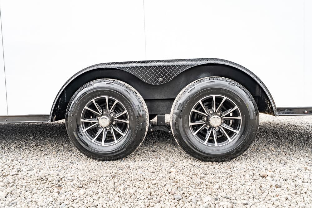 Twin-axle trailer wheels with black fenders on a white trailer, parked on gravel.