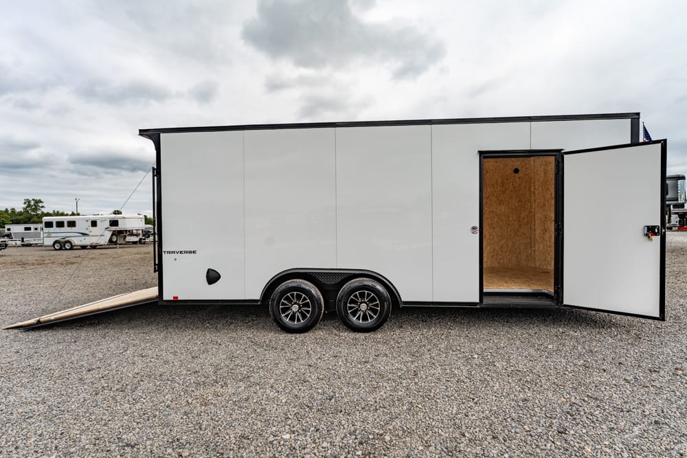 White enclosed cargo trailer with dual axles, ramp down, and side door open on a gravel lot; empty plywood interior visible.