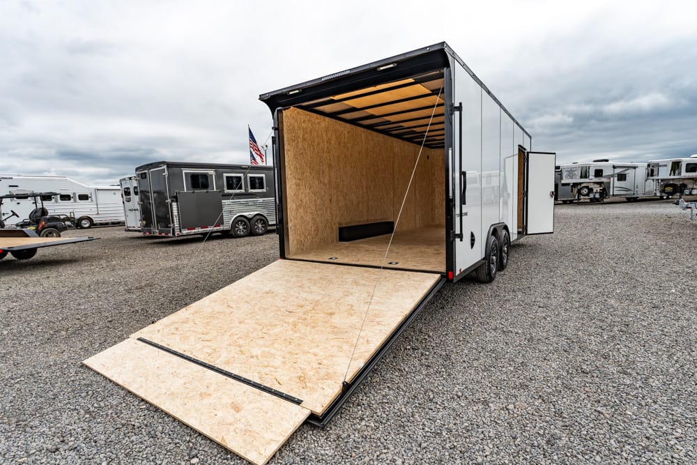 Open white enclosed cargo trailer with its plywood interior visible and a long wooden ramp extended onto a gravel lot.