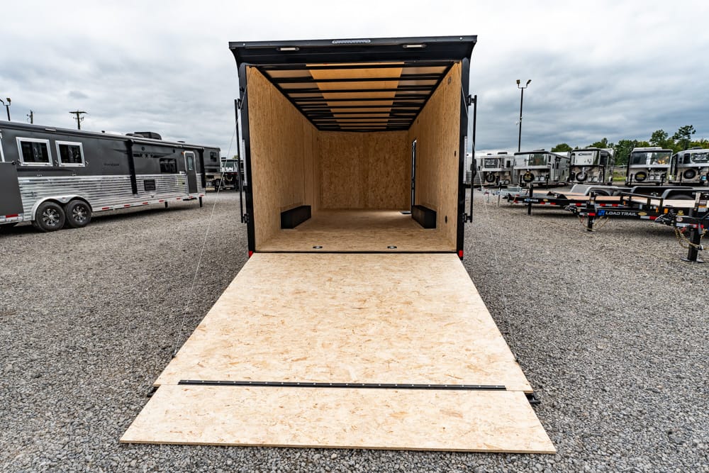 Open cargo trailer with ramp lowered onto gravel yard, showing plywood interior and surrounding trailers.