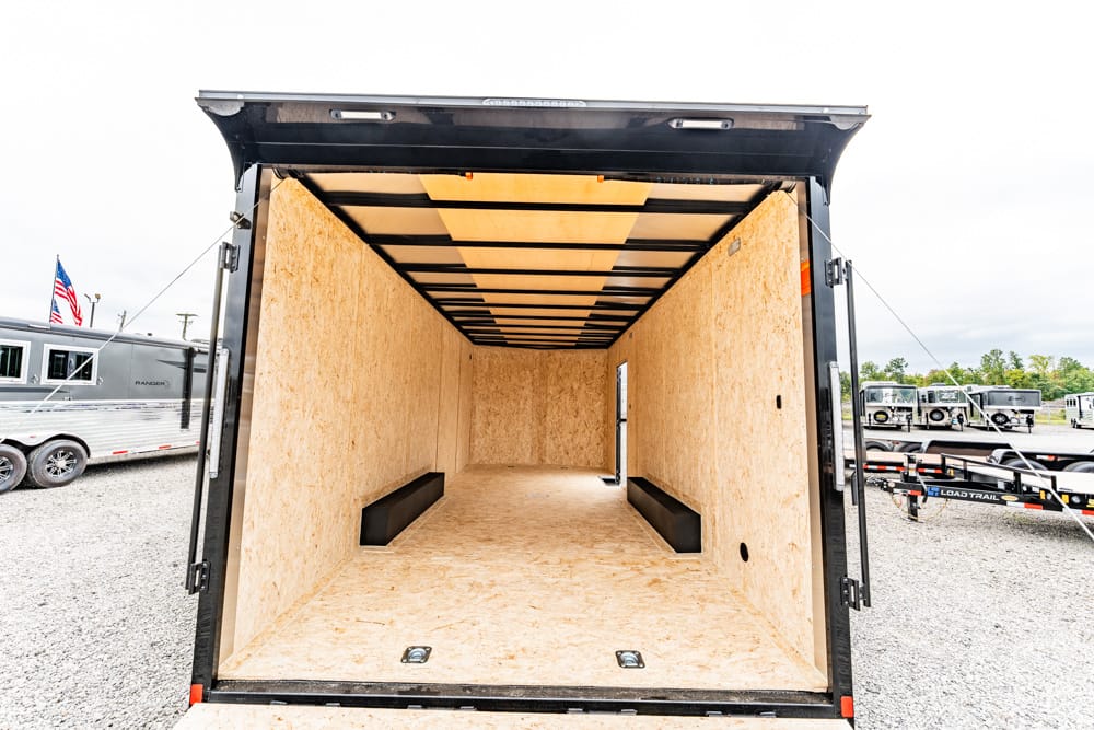 Open cargo trailer interior with unfinished plywood walls and ceiling, empty floor, and two black storage benches along the sides.