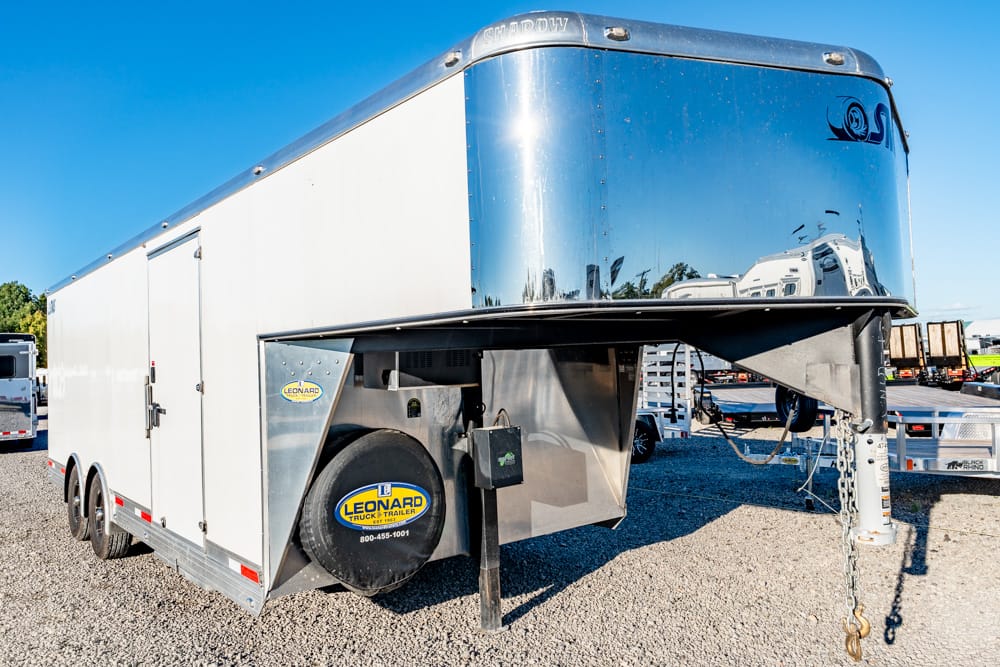 White enclosed gooseneck cargo trailer with a shiny chrome front, parked on a gravel lot; spare tire with Leonard logo visible at the front.