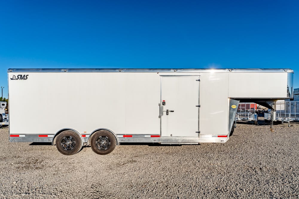 White enclosed utility trailer with dual rear axles parked on a gravel lot under a clear blue sky.