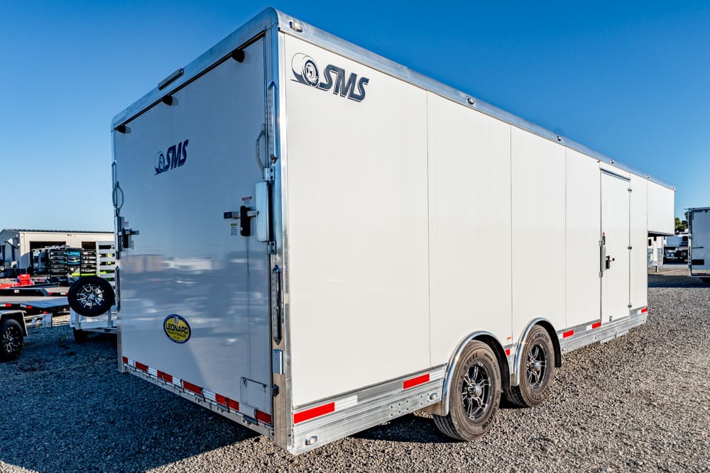 White enclosed cargo trailer with SMS branding parked on a gravel lot, two axles visible.