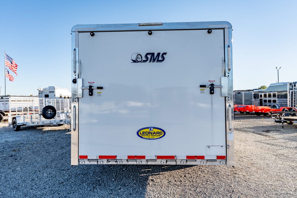 Rear view of a white cargo trailer in a gravel lot, featuring an SMS logo and a Leonard trailer sticker; American flag in the background.