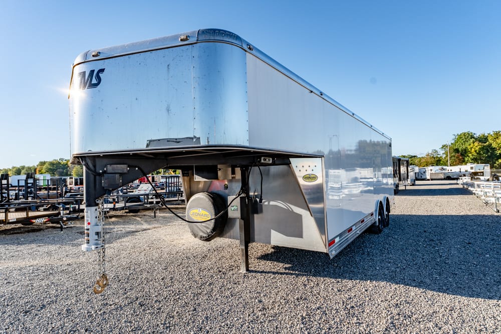 White enclosed cargo trailer on a gravel lot, front-right view with hitch and spare tire cover visible under a clear blue sky.